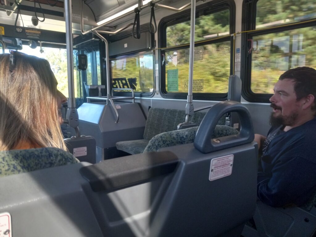 A white woman and white man talk with each other across the aisle on a bus.